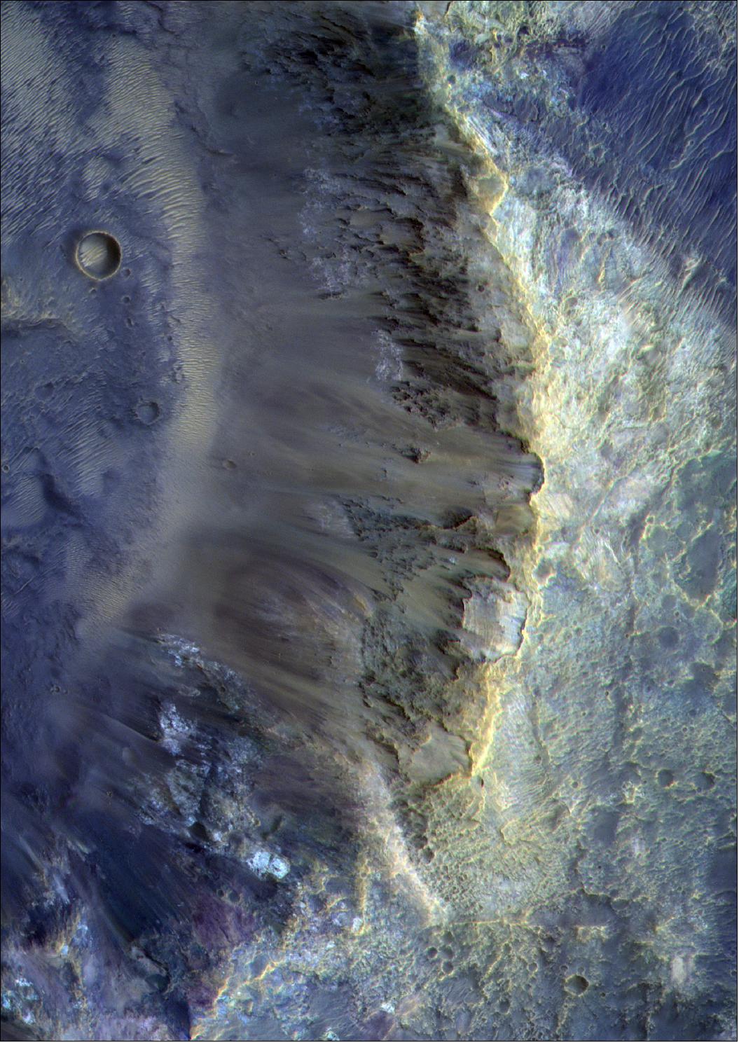 Figure 37: This image features the southeast wall of a small crater located a few hundred kilometers to the north of the giant Hellas impact basin on Mars. The complete crater itself is about 12 km in diameter; this image shows a 5 x 10 km area. When viewed with CaSSIS’ color filters, the image shows exceptional diversity in color. This diversity is related to the presence of various minerals that reflect light differently at different wavelengths. The light-toned deposits highlight the bedrock exposures of the area, which probably contain ancient clay-rich minerals that would have formed in the presence of water. Also visible are wind-blown sandy deposits that form ripples on the floor of the crater. Their distinctive tan color implies that they contain iron-oxide minerals. The image was featured by Science Advances online in February 2021 (image credit: ESA/Roscosmos/CaSSIS, CC BY-SA 3.0 IGO)