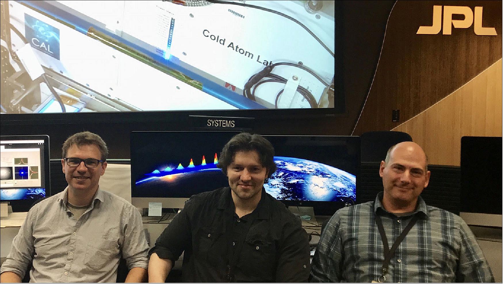 Figure 27: JPL scientists and members of the Cold Atom Lab's atomic physics team (left to right) David Aveline, Ethan Elliott and Jason Williams, shown here in the Earth Orbiting Missions Operation Center at JPL, where Cold Atom Lab (CAL) is remotely controlled and tuned. Displayed on the screen behind them is an image of CAL on the International Space Station. Aveline, Elliott and Williams were instrumental in producing the first ever Bose-Einstein condensates (BECs) in orbit with CAL (image credit: NASA/JPL-Caltech)